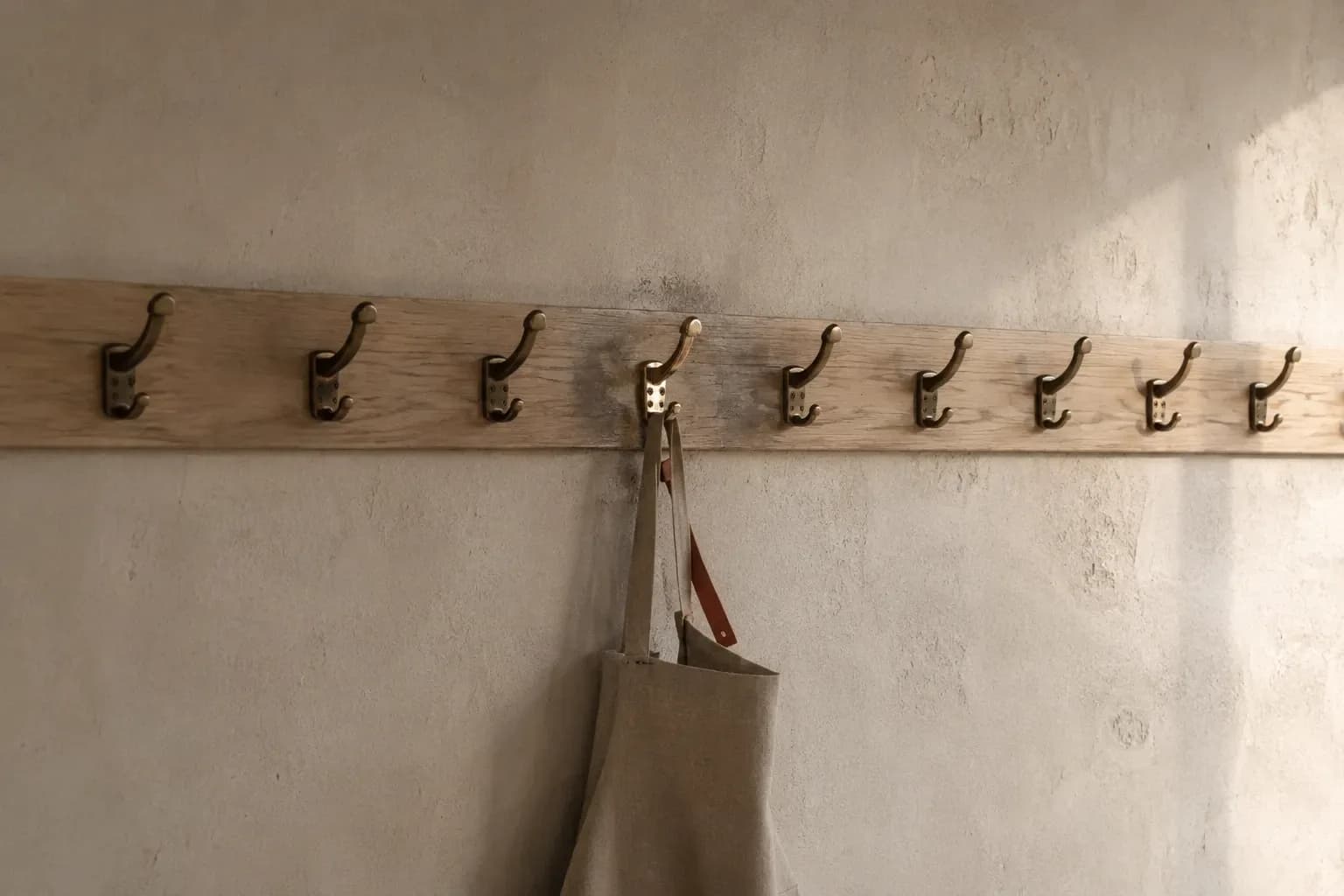 A long rail of brass hooks on a plain wall where one hook is polished bright and carries a hanging apron while the rest are dull and unused.