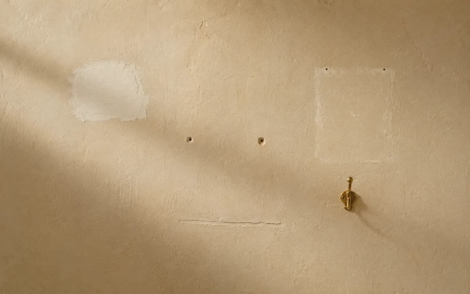 An aged plaster wall carrying a handful of small recent changes: one repainted patch, two filled screw holes, a new brass hook and a neatly repaired crack.