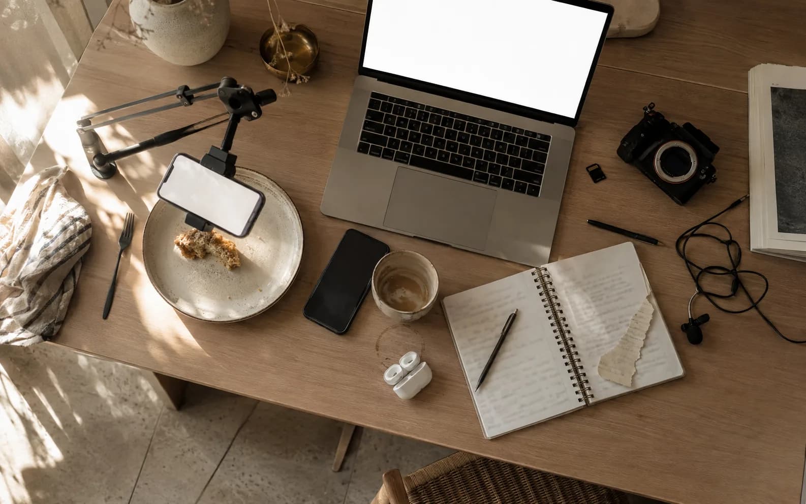 An overhead view of a working desk mid week: a phone clamped to an arm above it, a second phone face down, a laptop, a camera, a clip microphone and a cold coffee.