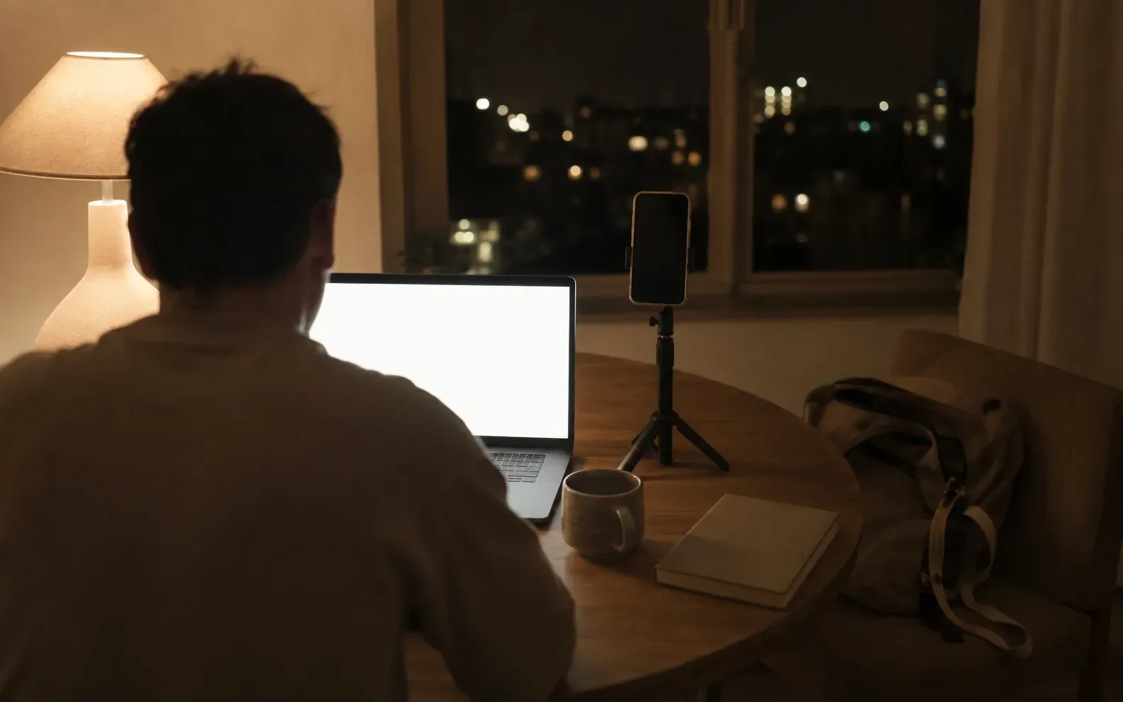 A kitchen table at night lit by one lamp, a laptop open and a phone on a small tripod beside it, with a work bag and lanyard dropped on the chair in the corner.