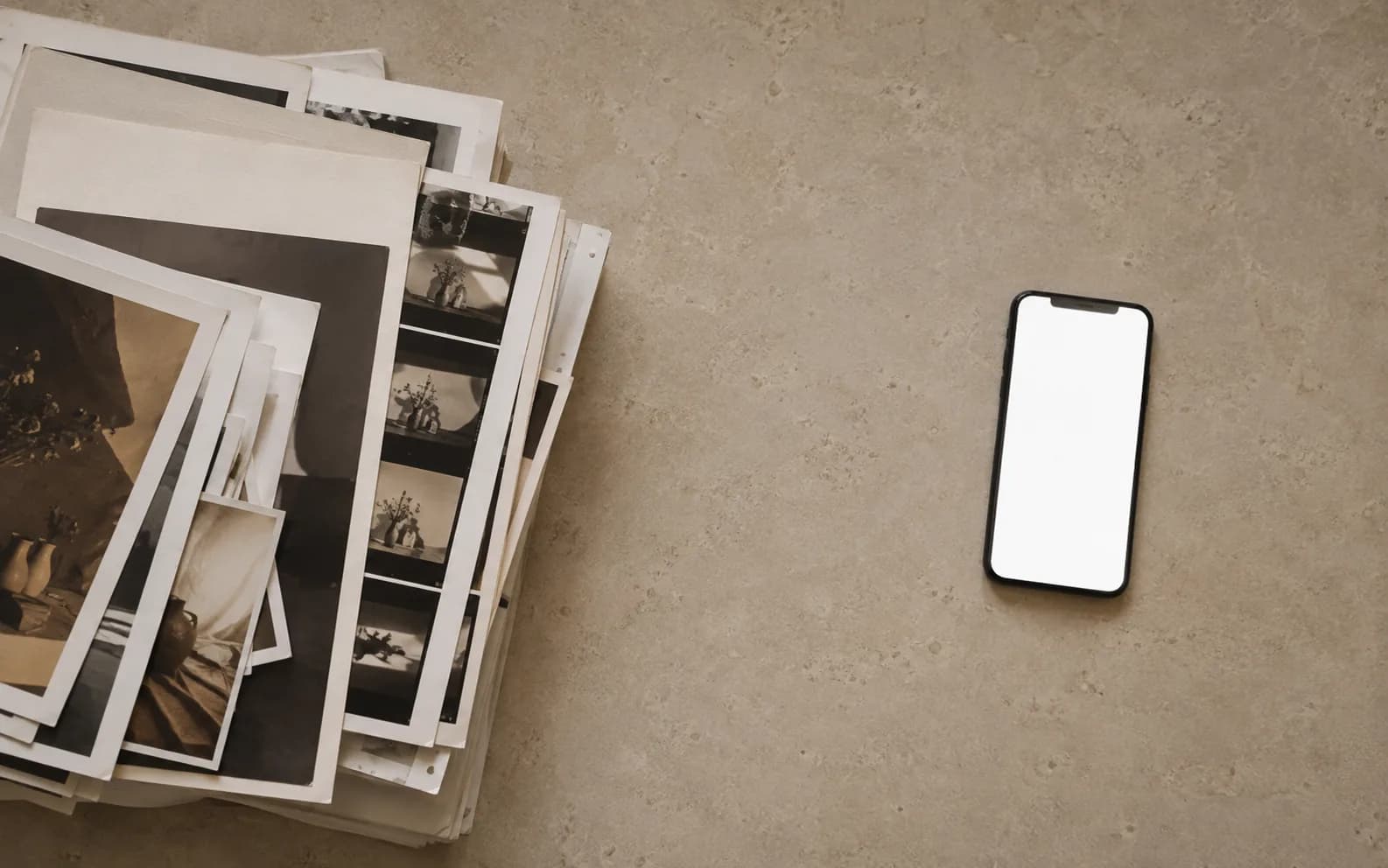 A thick stack of prints and contact sheets on one side of a surface and a phone with a blank bright screen on the other, with a clear empty gap between them.